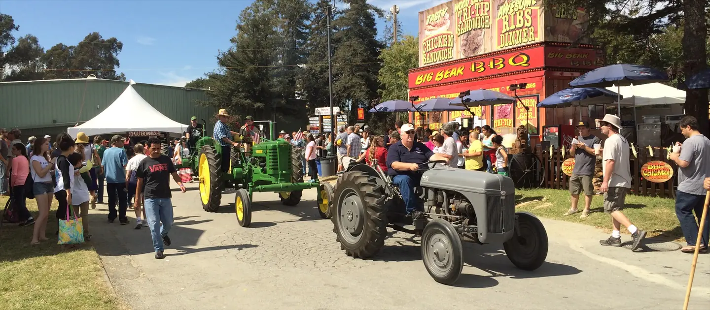 Two vintage tractors, a green John Deere and a gray Ford, in the tractor parade at the Santa Cruz County Fair, with fairgoers, a barbecue stand and redwoods behind