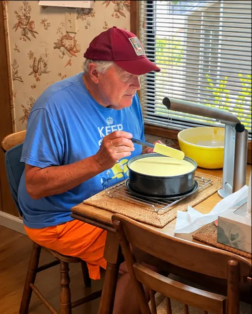 Garrett Wallace in a red cap at a wooden table, topping a cheesecake in its springform pan